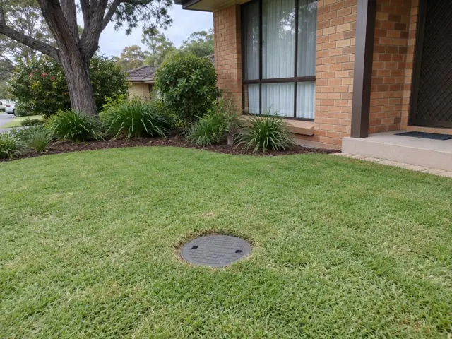 Circular capped sewer access cover set in a neatly kept front lawn beside a brick house in Frenchs Forest